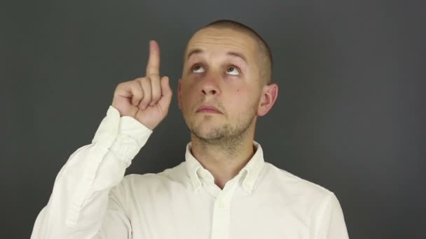 Lustig, stilig kille i vit skjorta visar pekfingret upp och nickar. Porträtt på en grå bakgrund. — Stockvideo