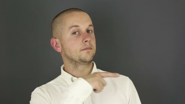 Zabawny, przystojny facet w białej koszuli pokazuje palec wskazujący z boku. Portret na szarym tle. — Wideo stockowe