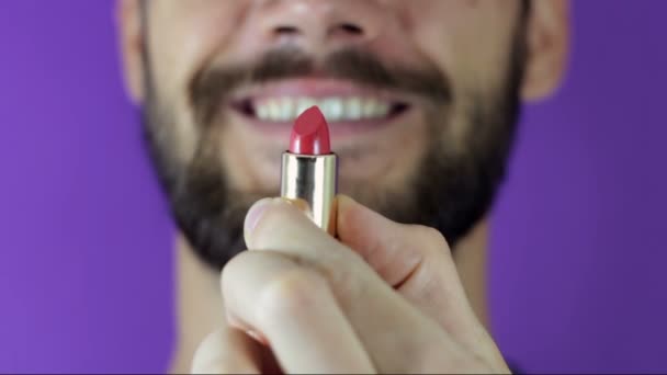 Ein junger Mann mit Bart blickt auf den roten Lippenstift und lächelt. Großaufnahme eines bärtigen Mannes. Er untersucht einen knallroten Lippenstift. Ein bärtiger Mann zieht Lippenstift hervor, schaut sie an und lächelt.. — Stockvideo