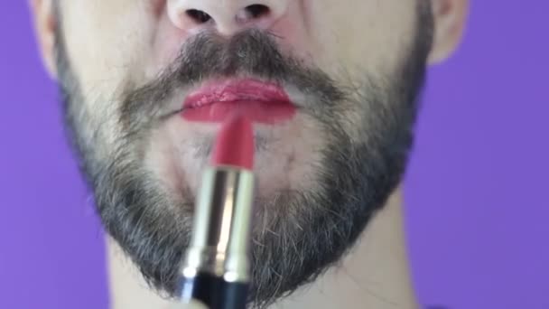 Un joven con barba se pinta los labios con lápiz labial rojo. Primer plano de un hombre barbudo, pintó los labios con lápiz labial brillante. Un barbudo se pone lápiz labial rojo en los labios, sonríe y se golpea los labios . — Vídeos de Stock