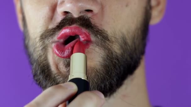 Un joven con barba se pinta los labios con lápiz labial rojo. Primer plano de un hombre barbudo, pintó los labios con lápiz labial brillante. Un barbudo se pone lápiz labial rojo en los labios, sonríe y se golpea los labios . — Vídeos de Stock