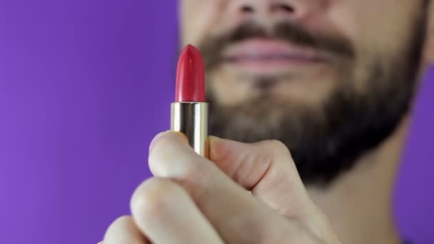 Ein junger Mann mit Bart blickt auf den roten Lippenstift und lächelt. Großaufnahme eines bärtigen Mannes. Er untersucht einen knallroten Lippenstift. Ein bärtiger Mann zieht Lippenstift hervor, schaut sie an und lächelt.. — Stockvideo