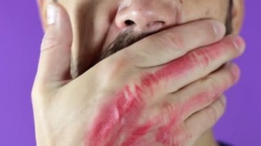 Sakallı genç bir adam yüzünde kırmızı rujla elini ovuşturuyor. Sakallı bir adam dudaklarına kırmızı ruj sürer, gülümser ve dudaklarına şaplak atar..