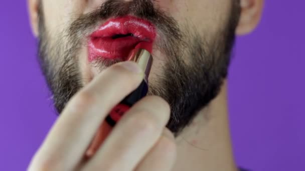 Un joven con barba se pinta los labios con lápiz labial rojo. Primer plano de un hombre barbudo, pintó los labios con lápiz labial brillante. Un barbudo se pone lápiz labial rojo en los labios, sonríe y se golpea los labios . — Vídeos de Stock