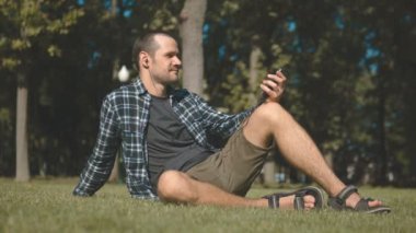Ekose gömlekli genç yakışıklı adam parkta çimlerin üzerinde otururken müzik dinliyor. Genç bir adam çimde otururken kulaklıkla müzik dinliyor..