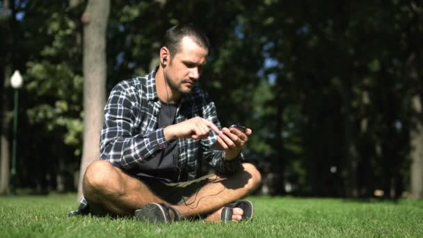公園の芝生の上に座っている間 遊んだシャツの若いハンサムな男は ソーシャルネットワークのフィードを通して葉 若い男は草の上に座っている間 彼のスマートフォンでソーシャルネットワークのフィードを読み取ります — ストック動画