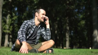 Genç bir adam çimlerin üzerinde otururken bir telefon görüşmesi yapıyor. Ekose gömlekli genç adam parkta çimlerin üzerinde otururken cep telefonuyla konuşuyor..