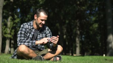Genç bir adam çimlerin üzerinde otururken akıllı telefonuyla sosyal ağların beslemesini okuyor. Ekose gömlekli genç adam parktaki çimlerin üzerinde otururken sosyal ağların yemlerini karıştırıyor..