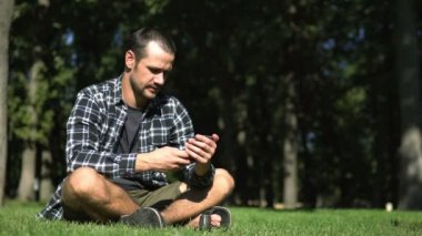 Genç bir adam çimlerin üzerinde otururken akıllı telefonuyla sosyal ağların beslemesini okuyor. Ekose gömlekli genç adam parktaki çimlerin üzerinde otururken sosyal ağların yemlerini karıştırıyor..