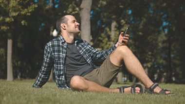 Ekose gömlekli genç yakışıklı adam parkta çimlerin üzerinde otururken müzik dinliyor. Genç bir adam çimlerin üzerinde otururken akıllı telefonuyla müzik dinliyor..