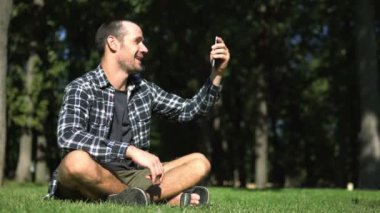 Genç bir adam çimlerin üzerinde otururken akıllı telefonuyla görüntülü konuşma yapıyor. Ekose gömlekli genç adam parkta çimlerin üzerinde otururken görüntülü konuşma yapıyor..