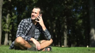 Genç bir adam çimlerin üzerinde otururken bir telefon görüşmesi yapıyor. Ekose gömlekli genç adam parkta çimlerin üzerinde otururken cep telefonuyla konuşuyor..