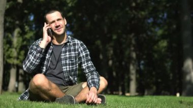 Genç bir adam çimlerin üzerinde otururken bir telefon görüşmesi yapıyor. Ekose gömlekli genç adam parkta çimlerin üzerinde otururken cep telefonuyla konuşuyor..