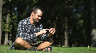 Genç bir adam çimlerin üzerinde otururken akıllı telefonuyla sosyal ağların beslemesini okuyor. Ekose gömlekli genç adam parktaki çimlerin üzerinde otururken sosyal ağların yemlerini karıştırıyor..