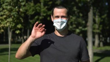 Genç ve çekici bir adam selamlaşmak için elini sallıyor ve koruyucu tıbbi maskesini çıkarıyor. Genç bir adam selamlaşmak için elini sallıyor koruyucu maskesini çıkarıyor ve rahatça iç çekiyor..