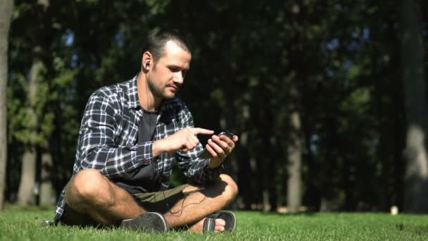 若い男が草の上に座っている間 スマートフォンでソーシャルネットワークのフィードを読み取ります 公園の草の上に座っている間 若いですハンサムな男で遊んだシャツの葉は ソーシャルネットワークのフィードを通して — ストック動画