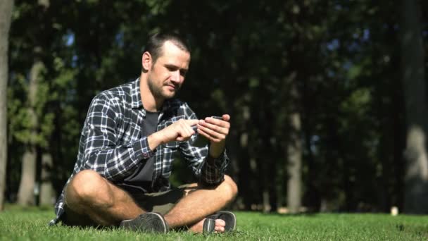 一个年轻人坐在草地上 一边用智能手机阅读社交网络的内容 年轻英俊的小伙子穿着格子呢衬衫 坐在公园的草地上 靠社交网络的支持度日 — 图库视频影像