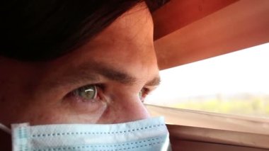 Tıbbi maske takmış genç yakışıklı bir adam pencere perdelerinden içeri bakıyor. Tıbbi maskeli genç bir adam pencereden bakar, kendini izole eder..