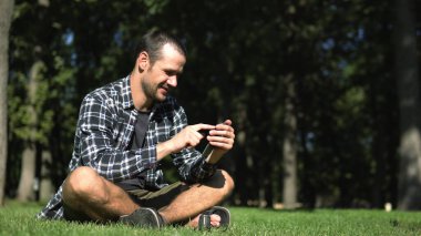 Ekose gömlekli genç adam parktaki çimlerin üzerinde otururken sosyal ağların beslemelerini karıştırıyor. Genç bir adam akıllı telefonunda otururken sosyal ağların beslemesini okuyor.