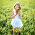Cute little child in white dress posing in green field and looking at camera