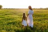 Mutter und Tochter halten sich an den Händen und spazieren an einer grünen Wiese mit Sonnenuntergang im Hintergrund vorbei