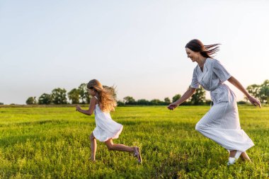 active mother and daughter in white dresses running in green meadow clipart