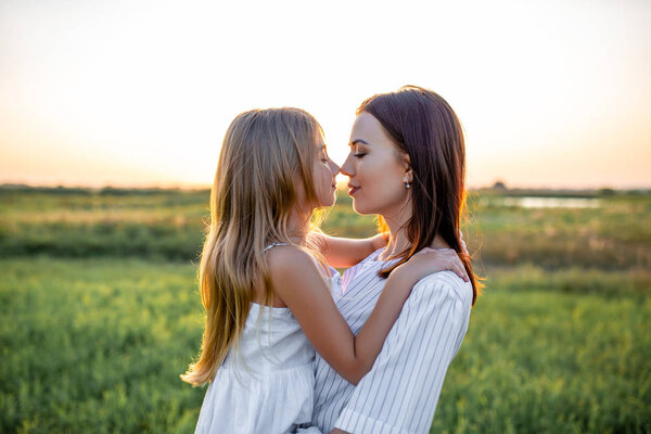 close-up portrait of beautiful mother and daughter embracing and touching noses in green field on sunset