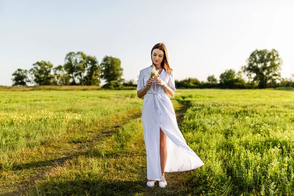 有魅力的年轻妇女在白色礼服与田野花花束在绿色领域 图库照片