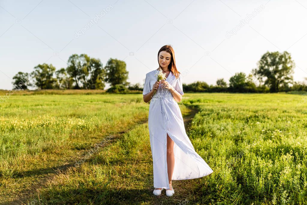 attractive young woman in white dress with field flowers bouquet on green field