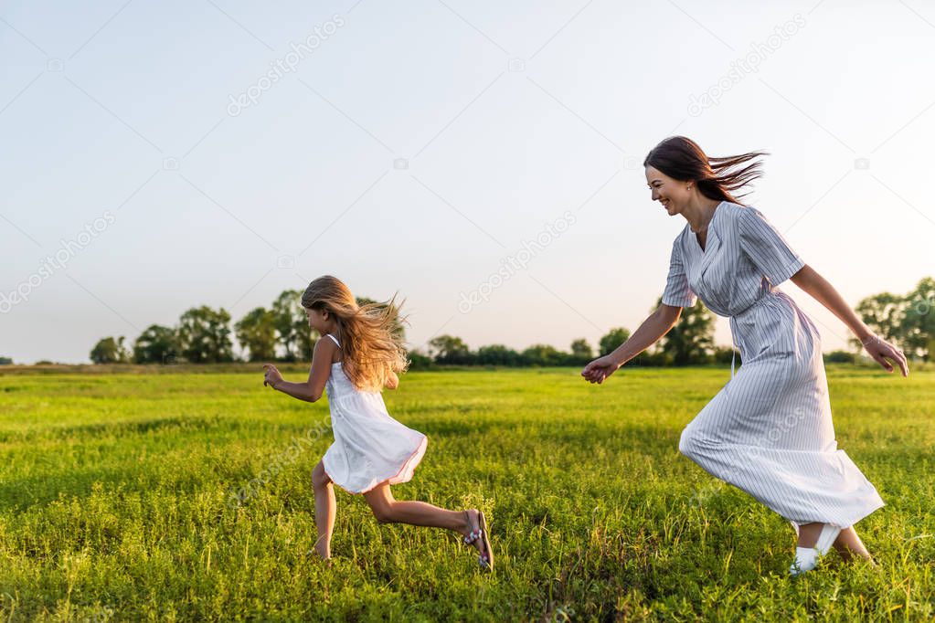 active mother and daughter in white dresses running in green meadow