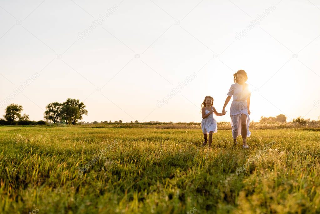 mother and daughter walking in green meadow with sunset on background