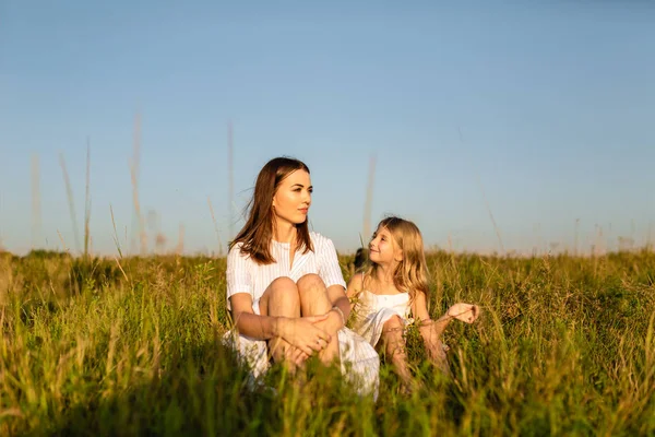 Madre e hija sentadas sobre hierba verde al atardecer y charlando - foto de stock