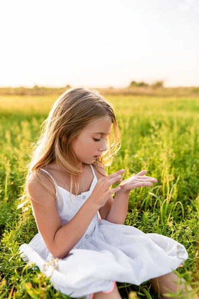 Чарівна маленька дитина сидить на зеленій траві і дивиться на руку під променями заходу сонця — стокове фото