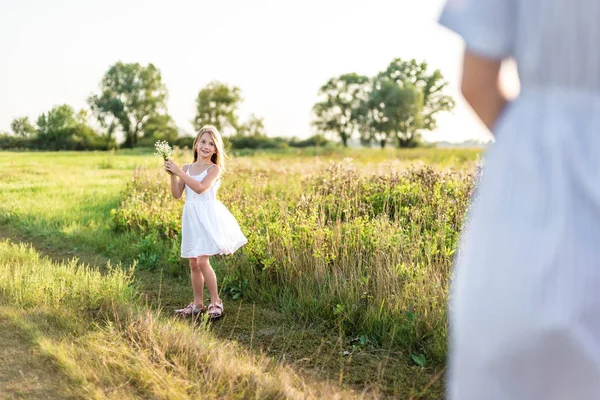 Menina feliz coletando flores no prado verde e olhando para a mãe — Fotografia de Stock