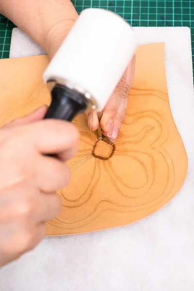 workshop of making the carved leather bag - craftsman stamps the drawing of flower on surface of rough vegetable tanned leather with Stamping tool and mallet