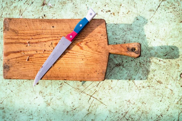 top view of steel knife with a red-blue-white handle on a dirty wooden cutting board on a outdoor messy table