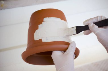 Beyaz eldivenli genç bir kadının elleri seramik bir bahçe kabını boyar. Bahçede çalışmak, çiçek açan sardunyalar, yeşil yapraklar ve sulu etlerin arka planına karşı çömlek dikim.