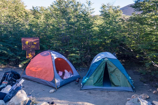 Two opened and messy camping tents and chaotic gear at a remote campsite in the wilderness , next to a forest in the morning or the afternoon, with a ilegible sign