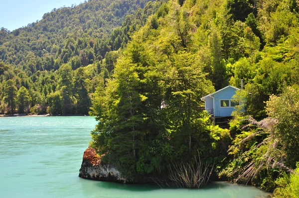 Idyllic, heavenly, beautiful light blue small house surrounded by nature, inside a forest and next to a river with cold turquoise waters, near the mountains, in Carretera Austral, Patagonia, Chile