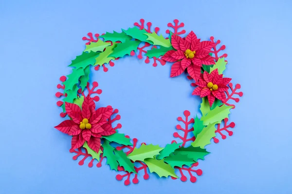 Christmas wreath of paper flowers poinsettia. Favorite hobby is manual work. Green holly and red berries. Blue background.