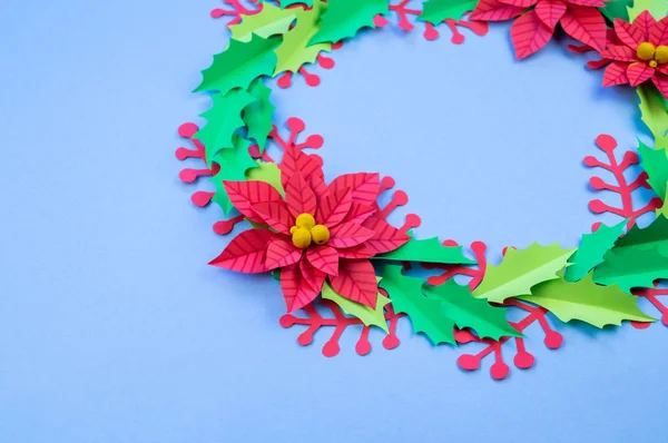 Christmas wreath of paper flowers poinsettia. Favorite hobby is manual work. Green holly and red berries. Blue background.