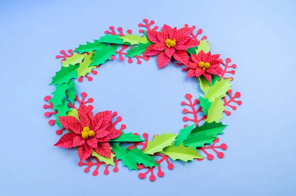 Christmas wreath of paper flowers poinsettia. Favorite hobby is manual work. Green holly and red berries. Blue background.