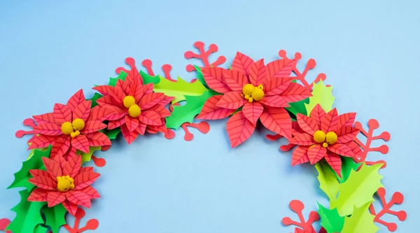 Christmas wreath of paper flowers poinsettia. Favorite hobby is manual work. Green holly and red berries. Blue background.