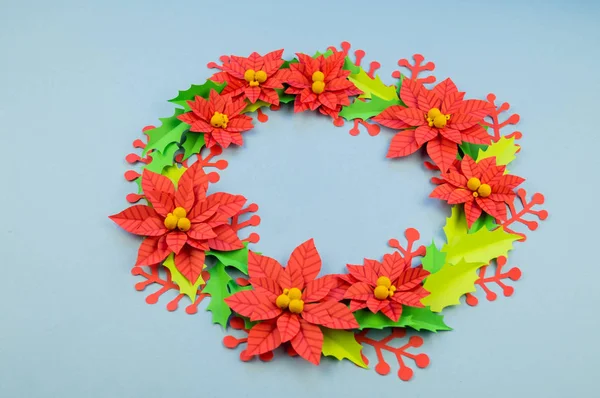 Christmas wreath of paper flowers poinsettia. Favorite hobby is manual work. Green holly and red berries. Blue background.