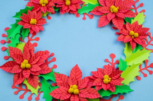 Christmas wreath of paper flowers poinsettia. Favorite hobby is manual work. Green holly and red berries. Blue background.