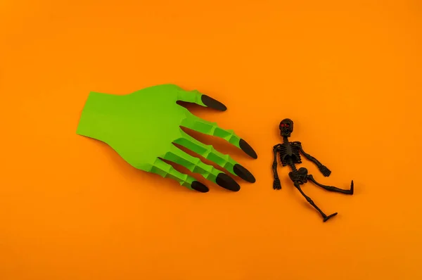 Orange background with collection of Halloween objects origami overhead view. Green monster hand. Feared party decor. Handwork.