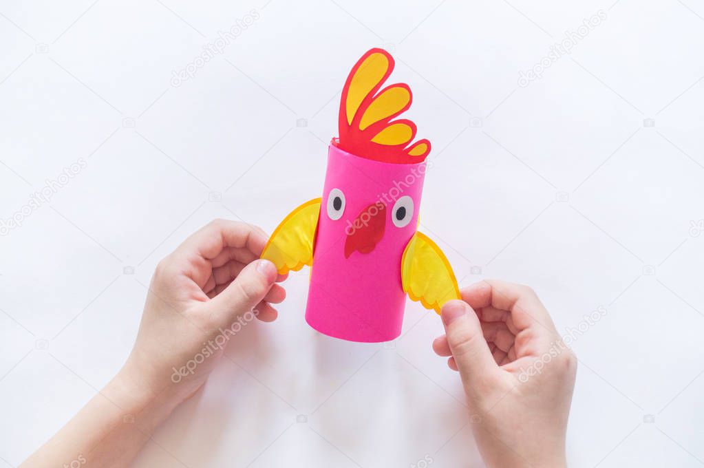 The child makes a craft from cardboard. Paper parrot pink. Hands girls.