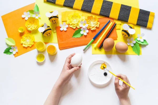 Gyermek kezek lány fest egy húsvéti tojást. Tojás egy sárga méh. Fehér háttér. Anyag kreativitásért, — Stock Fotó