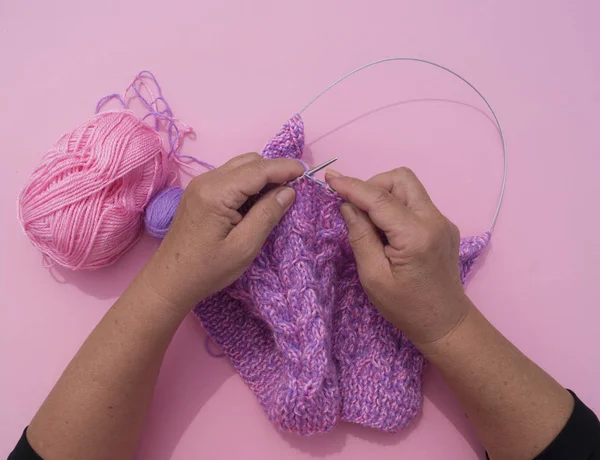 As mãos da mulher tricotam um gorro de fio roxo. Fundo rosa — Fotografia de Stock