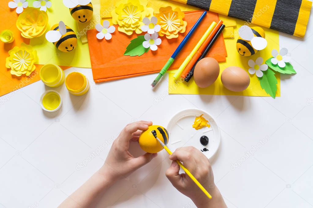 Child hands girl paints an Easter egg. Egg a yellow bee. White background. Material for creativity,